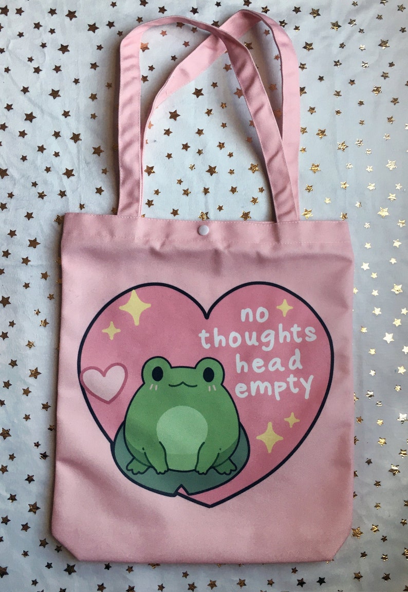 May include: Pink canvas tote bag with a green frog and a heart design. The text on the bag reads "no thoughts head empty".