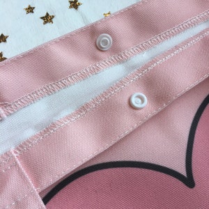May include: Close-up of a pink canvas tote bag with white snaps. The bag has a black heart design and a white and yellow star pattern.