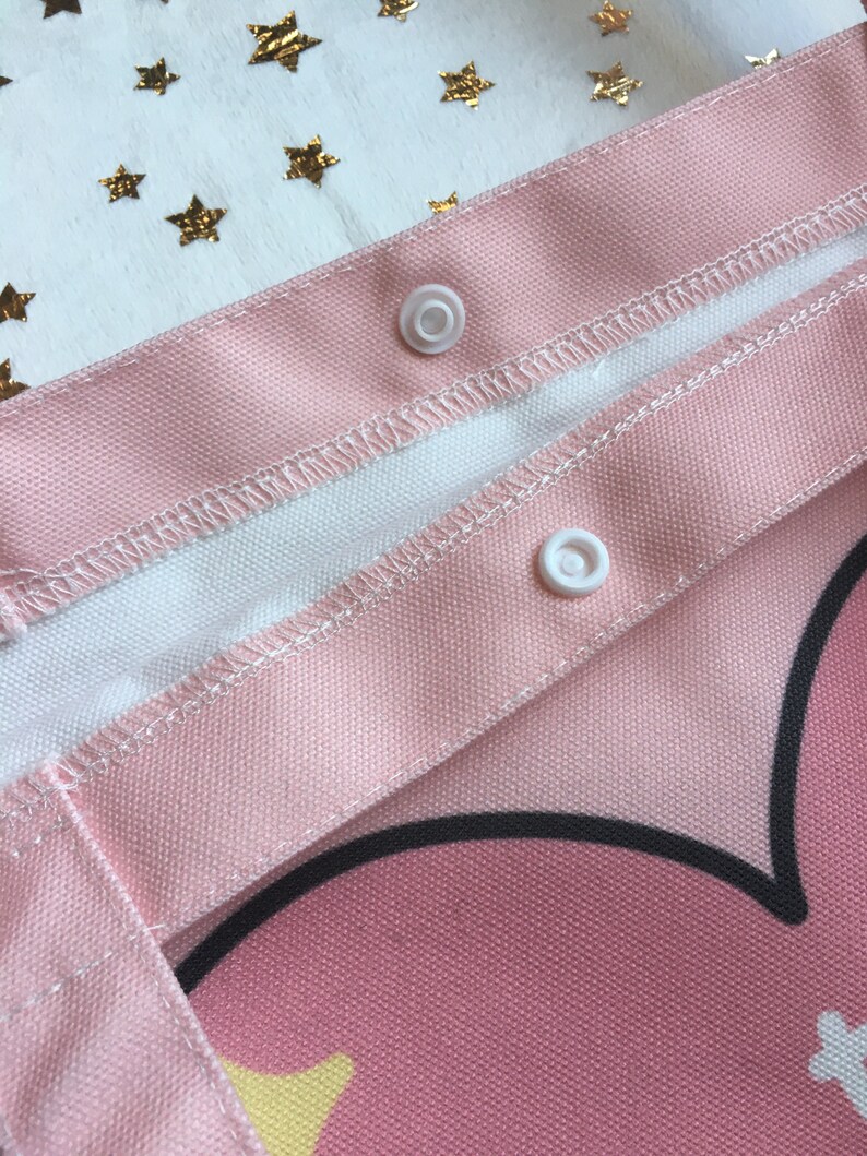 May include: Close-up of a pink canvas tote bag with white snaps. The bag has a black heart design and a white and yellow star pattern.