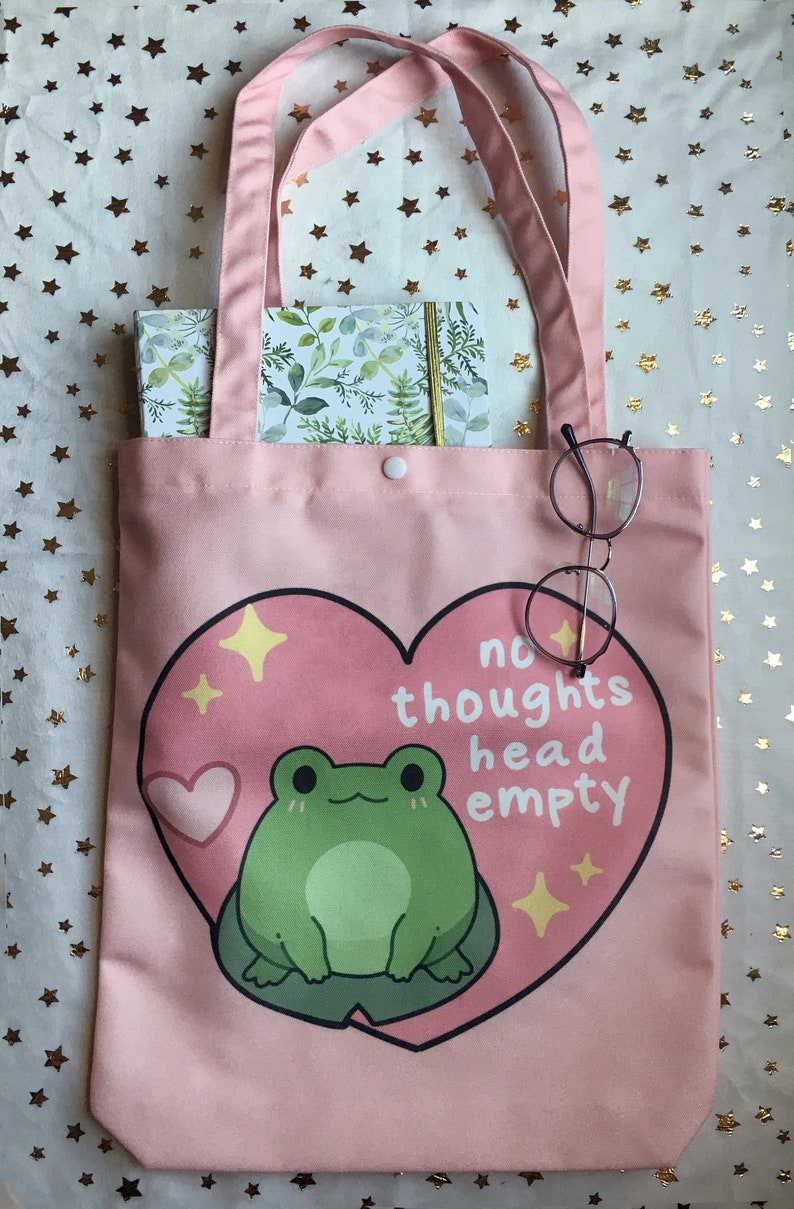 May include: A pink canvas tote bag with a white button closure. The bag features a heart-shaped design with a green frog inside and the text "no thoughts head empty".