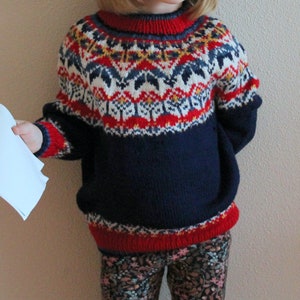 Puede incluir: Un niño o niña lleva un suéter de punto azul, rojo, blanco y amarillo con un patrón escandinavo tradicional. El suéter tiene cuello redondo y mangas largas.