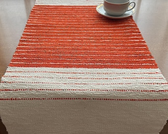 May include: A woven table runner with a design of alternating white and orange stripes. A white teacup and saucer with a gold rim sit on the runner, which is placed on a wooden table. The runner is approximately 60 inches long.