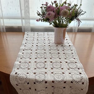 May include: A white, crocheted table runner with a floral pattern is displayed on a wooden table. A vase with pink and purple flowers sits on the table. The table has a semi-circular shape.