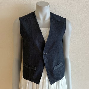 以下が含まれることがあります： ボタン留めとポケットが2つ付いた、濃い青色のデニムベスト。ベストは袖なしで、尖った襟が付いています。