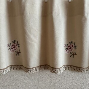 May include: A white valance with a brown lace trim and embroidered floral designs. The valance is hung on a curtain rod with white clips.