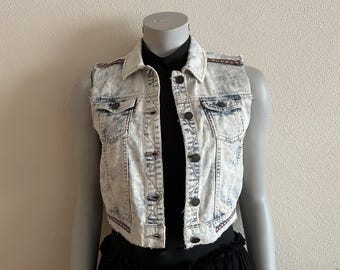 May include: A sleeveless denim vest with a light blue and white acid wash. The vest has a collared neckline, button-down front, and two chest pockets. The shoulders and hem are accented with a decorative trim.