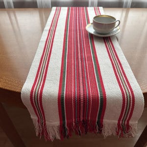 May include: A striped table runner in white, red, green, and pink, with a fringed edge. The runner is placed on a wooden table. A teacup and saucer with a gold rim sit on the runner. The table runner is approximately 150 cm long.