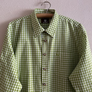 Può includere: Una camicia a bottoni a quadri verdi e bianchi con una sola tasca sul lato sinistro. La camicia ha un colletto e maniche lunghe.