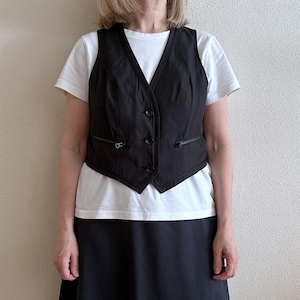 May include: A black sleeveless vest with a zipper closure and pockets. The vest is worn over a white t-shirt and a black skirt.