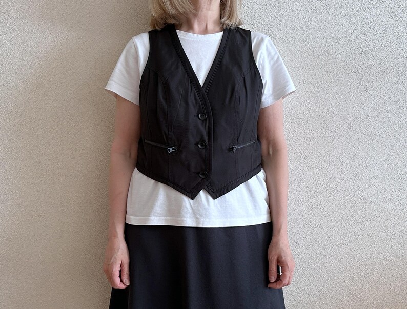 May include: A black sleeveless vest with a zipper closure and pockets. The vest is worn over a white t-shirt and a black skirt.