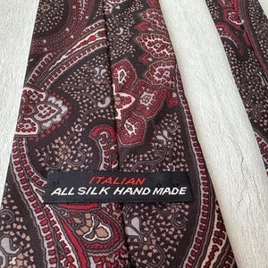 Op de afbeelding: Close-up van een zijden stropdas met een paisley patroon in bruin, rood en cr&egrave;me tinten. De stropdas heeft een label met de tekst "ITALIAN ALL SILK HAND MADE". Een klassiek accessoire.
