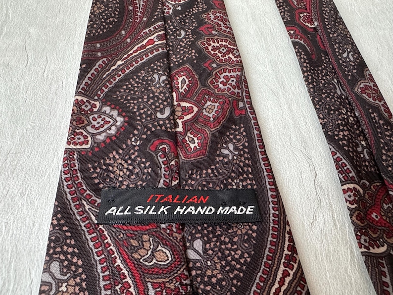 Op de afbeelding: Close-up van een zijden stropdas met een paisley patroon in bruin, rood en cr&egrave;me tinten. De stropdas heeft een label met de tekst "ITALIAN ALL SILK HAND MADE". Een klassiek accessoire.
