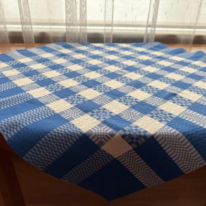 May include: A square tablecloth featuring a blue and white checkered pattern with intricate woven geometric details within the squares. The fabric displays a textured weave throughout, creating a repeating grid design across the entire surface.
