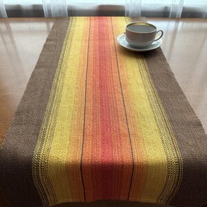 Può includere: Runner da tavolo tessuto con una sfumatura di colori che include marrone, giallo, arancione e rosso. Una tazza da tè bianca con bordo dorato su un piattino. Il runner è su un tavolo di legno.