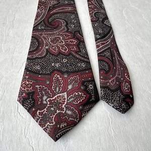 Op de afbeelding: Een stropdas met paisley-patroon in zwart, bordeauxrood en cr&egrave;me. Het paisley-patroon is gedetailleerd en complex, met een zich herhalend bloemmotief. De stropdas heeft een klassieke vorm en is gemaakt van gladde stof.