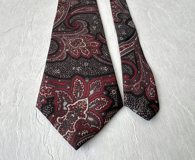 Op de afbeelding: Een stropdas met paisley-patroon in zwart, bordeauxrood en cr&egrave;me. Het paisley-patroon is gedetailleerd en complex, met een zich herhalend bloemmotief. De stropdas heeft een klassieke vorm en is gemaakt van gladde stof.