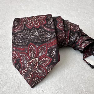 Op de afbeelding: Een stropdas met patroon met een paisley-ontwerp in tinten bordeauxrood, rood en zwart. De stropdas is opgevouwen en ligt op een getextureerd, lichtgekleurd oppervlak. Het ingewikkelde bloemenpatroon is zichtbaar.
