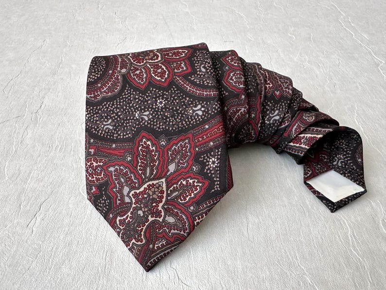 Op de afbeelding: Een stropdas met patroon met een paisley-ontwerp in tinten bordeauxrood, rood en zwart. De stropdas is opgevouwen en ligt op een getextureerd, lichtgekleurd oppervlak. Het ingewikkelde bloemenpatroon is zichtbaar.