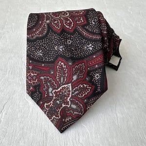 Op de afbeelding: Een patroon stropdas met een paisley design in bordeaux, zwart en grijs. De stropdas is geknoopt en toont de ingewikkelde bloemen- en geometrische patronen.