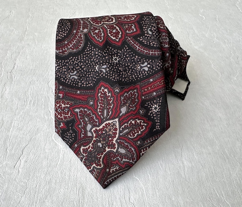 Op de afbeelding: Een patroon stropdas met een paisley design in bordeaux, zwart en grijs. De stropdas is geknoopt en toont de ingewikkelde bloemen- en geometrische patronen.