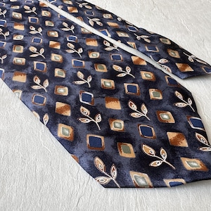 Könnte beinhalten: Eine dunkelblaue Krawatte mit einem sich wiederholenden Muster aus kleinen Quadraten in Braun-, Hellblau- und Beigetönen, durchsetzt mit weiß umrandeten Blattmotiven. Die Krawatte hat eine glatte, leicht reflektierende Oberfläche.