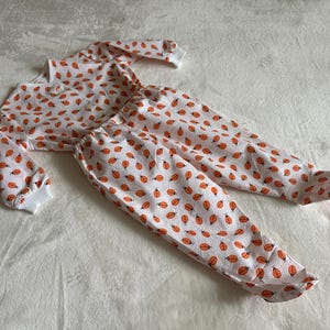Könnte beinhalten: Weißes Pyjama-Set mit einem Muster aus orangefarbenen Marienkäfern. Das langärmlige Oberteil hat eine Knopfleiste und weiße gerippte Bündchen. Die passende Hose hat einen elastischen Bund und Fußenden.