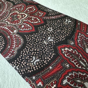 Op de afbeelding: Een close-up van een stropdas met patroon. De stropdas heeft een paisley-ontwerp in tinten rood, zwart en beige. Het ingewikkelde patroon is zichtbaar over de stof en cre&euml;ert een gedetailleerde en getextureerde uitstraling. De stropdas is schuin geplaatst en toont het volledige ontwerp.