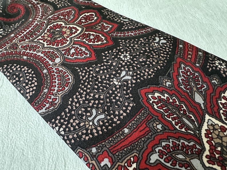 Op de afbeelding: Een close-up van een stropdas met patroon. De stropdas heeft een paisley-ontwerp in tinten rood, zwart en beige. Het ingewikkelde patroon is zichtbaar over de stof en cre&euml;ert een gedetailleerde en getextureerde uitstraling. De stropdas is schuin geplaatst en toont het volledige ontwerp.