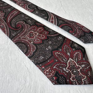 Op de afbeelding: Een stropdas met patroon met een paisley-ontwerp in zwart, rood en grijs. De stropdas is gemaakt van een gladde stof en heeft een klassieke vorm. Het ingewikkelde patroon is over de gehele lengte van de stropdas zichtbaar.