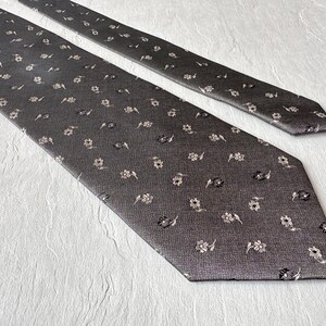 Può includere: Una cravatta grigio antracite con un motivo ripetuto di piccoli disegni floreali bianchi sporchi e neri. La cravatta è realizzata in un tessuto strutturato e viene presentata su una superficie bianca.