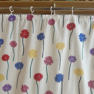 Könnte beinhalten: Ein weißer Vorhang mit einem sich wiederholenden Muster aus stilisierten roten, gelben, blauen und violetten Blumen mit grünen Stielen. Der Stoff hängt an einer silbernen Metallstange an weißen Kunststoffringen. Der Text am rechten Rand lautet ASTER design 1976.