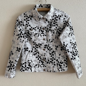 May include: White denim jacket with a black floral pattern. The jacket has a button-down front, two chest pockets, and long sleeves. The jacket is hanging on a wooden hanger.