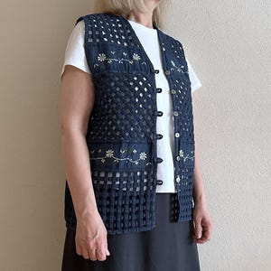 Puede incluir: Un chaleco vaquero azul con un diseño de crochet y bordado floral. El chaleco tiene cierre de botones y es sin mangas.