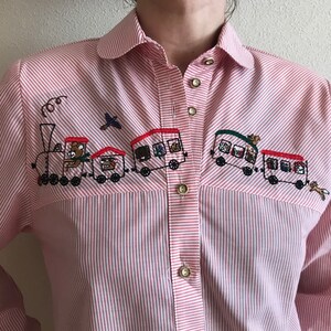 May include: A pink and white striped button-down shirt with an embroidered train design. The train is made up of several carriages with teddy bears and other animals inside. The shirt has a collar and long sleeves.