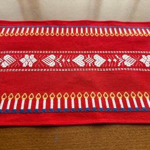 Può includere: Tessuto rosso con motivi ricamati bianchi, gialli e blu. Il design include file di candele stilizzate e motivi floreali o a forma di cuore. Probabilmente un runner da tavolo o un tessuto decorativo.