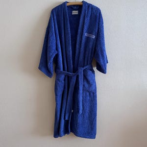 May include: A royal blue terry cloth robe hanging on a wooden hanger. The robe has a shawl collar, long sleeves, and a matching belt. A small label is visible on the chest. The robe is made of a thick, absorbent material.
