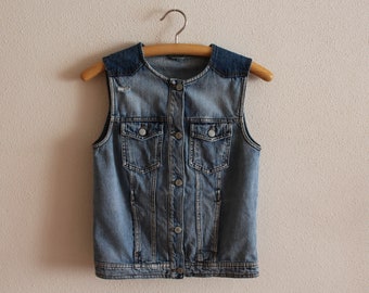 May include: A light blue denim vest with a button closure and two front pockets. The vest has a dark blue denim collar and is hanging on a wooden hanger.