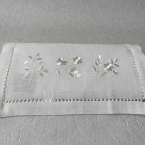 May include: White rectangular linen cloth with embroidered floral designs. The embroidery features three sets of flowers and leaves in shades of gray and white. The edges are finished with a decorative stitch.