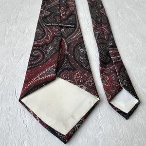 Op de afbeelding: Een zijden stropdas met een paisley patroon in tinten bordeaux, zwart en grijs. De stropdas is handgemaakt en heeft een witte voering. De stropdas wordt gepresenteerd op een witte ondergrond.