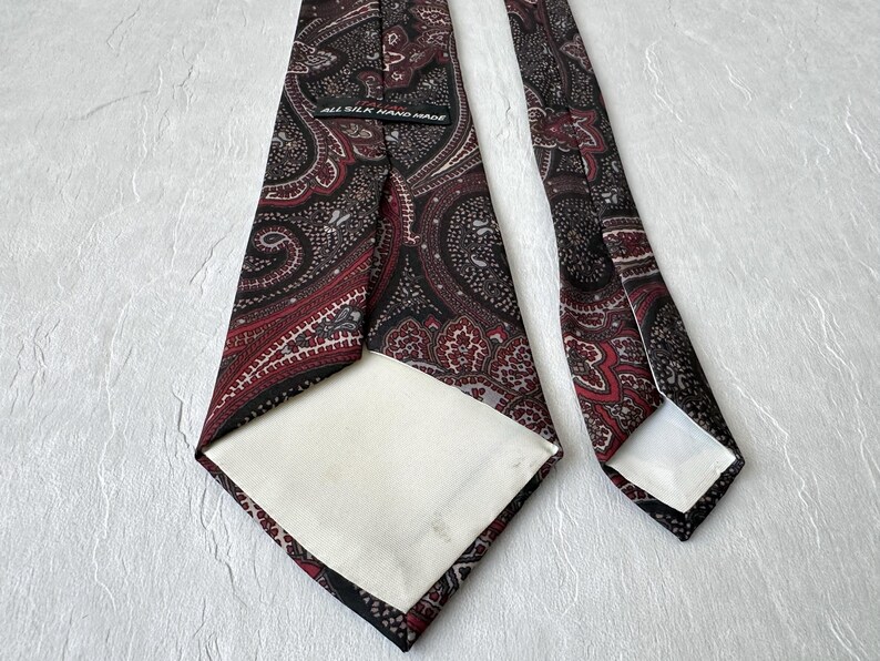 Op de afbeelding: Een zijden stropdas met een paisley patroon in tinten bordeaux, zwart en grijs. De stropdas is handgemaakt en heeft een witte voering. De stropdas wordt gepresenteerd op een witte ondergrond.