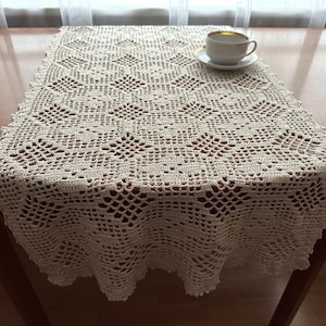 Könnte beinhalten: Cremefarbener, gehäkelter Tischläufer mit geometrischem Muster. Der Läufer liegt auf einem Holztisch, und eine weiße Tasse und Untertasse mit Goldrand stehen darauf. Das filigrane Lochmuster ist sichtbar.
