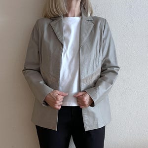 Pode incluir: Um blazer cinza claro com tecido ligeiramente texturizado. O blazer tem um colarinho de entalhe, mangas compridas e um fecho de botão único. O blazer tem um detalhe de design subtil com uma guarnição de cor castanho claro contrastante ao longo das bordas das lapelas e dos bolsos.