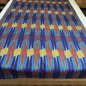 Può includere: Un tessuto grande e colorato con un motivo geometrico ripetuto. Il design presenta una combinazione di forme blu, arancioni e gialle, creando un tessuto vibrante e accattivante. Il tessuto è steso, mostrando il suo motivo completo.