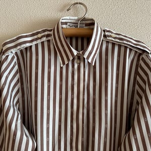 Puede incluir: Camisa a rayas marrones y blancas con cuello y mangas largas. La camisa tiene pequeños patrones en forma de cruz entre las rayas. La camisa está colgada en una percha de madera.
