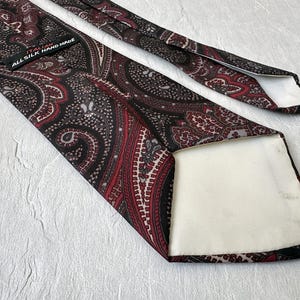 Op de afbeelding: Een zijden stropdas met een paisley patroon in bordeaux, zwart en grijs. De stropdas heeft een gedetailleerd ontwerp en de tekst "ALL SILK HAND MADE". Een klassiek accessoire.
