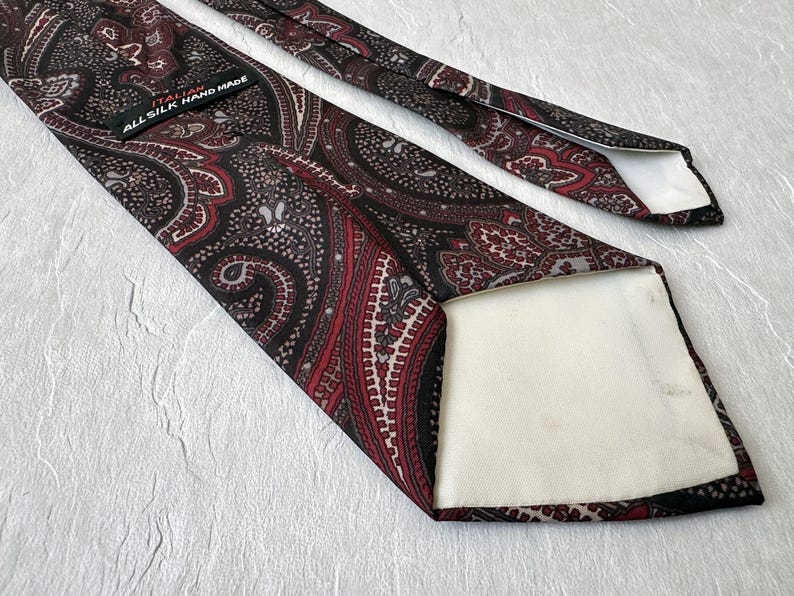 Op de afbeelding: Een zijden stropdas met een paisley patroon in bordeaux, zwart en grijs. De stropdas heeft een gedetailleerd ontwerp en de tekst "ALL SILK HAND MADE". Een klassiek accessoire.