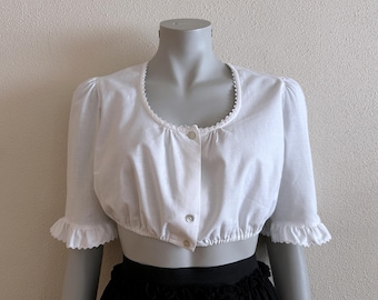 May include: A white, cropped blouse with a rounded neckline and short sleeves. The neckline and sleeve cuffs are trimmed with a delicate, scalloped edge. The blouse fastens with small, round buttons down the front. The garment is displayed on a mannequin.