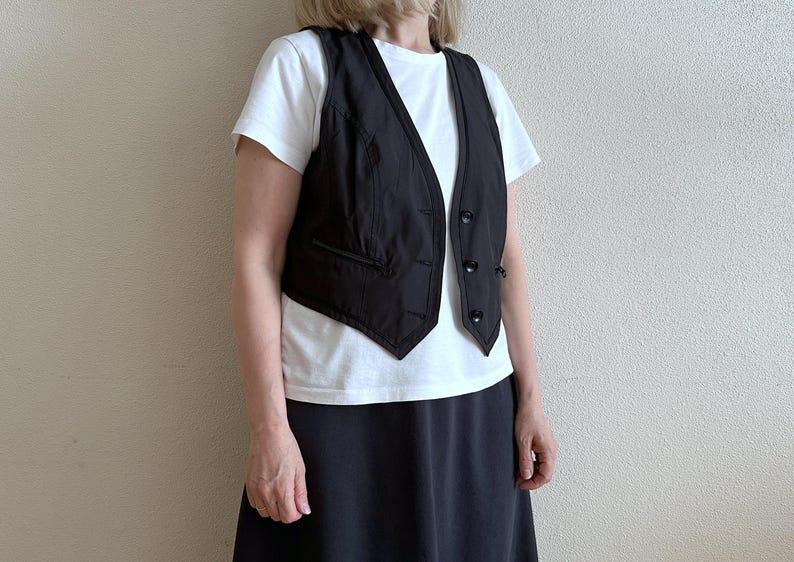 May include: A black vest with a white shirt and a black skirt. The vest has a button closure and a pocket on the left side.