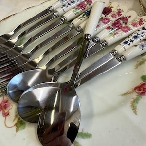 May include: A set of stainless steel cutlery featuring floral patterned handles. The set includes forks and spoons, arranged on a decorative plate with a floral design. The handles have a white base with pink, blue, and white flower patterns.