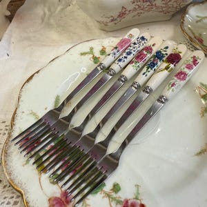 May include: A set of six vintage forks with floral patterned handles. The handles display various floral designs in pink, blue, and red. The forks are arranged on a decorative plate with a gold rim and floral accents.